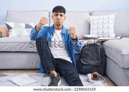 Young hispanic man sitting on the floor studying for university angry and mad raising fists frustrated and furious while shouting with anger. rage and aggressive concept. 