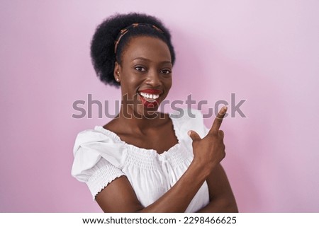 African woman with curly hair standing over pink background with a big smile on face, pointing with hand finger to the side looking at the camera. 