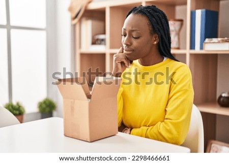 Beautiful black woman opening cardboard box thinking concentrated about doubt with finger on chin and looking up wondering 