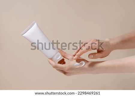 Fragile hands of a woman hold a white mockup tube of facial cream and apply moisturizer to her skin on a beige isolated background. Image for your design. Royalty-Free Stock Photo #2298469261