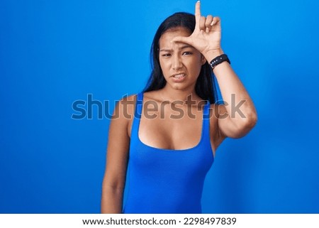 Hispanic woman standing over blue background making fun of people with fingers on forehead doing loser gesture mocking and insulting. 
