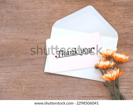 White greeting card with the words written "Thank you" and red hreart shape on a light green envelope and orange chrysanthemum blooming on rustic wooden background.
