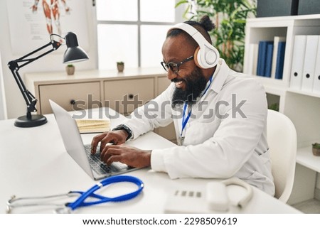 Young african american man wearing doctor uniform using laptop and headphones working at clinic