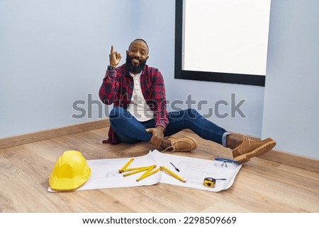 African american man sitting on the floor at new home looking at blueprints pointing finger up with successful idea. exited and happy. number one. 