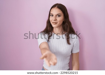 Young hispanic girl standing over pink background smiling cheerful offering palm hand giving assistance and acceptance. 