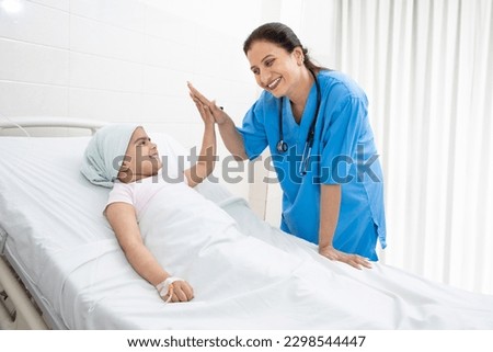 Happy indian woman nurse or medical staff give a high five to little girl cancer patient lying on hospital bed undergoing course of chemotherapy. Royalty-Free Stock Photo #2298544447