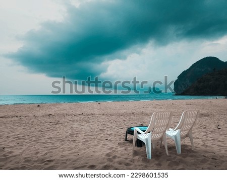 A deserted Beach in Thailand with Two White Plastic Garden Beach Chairs on the Sand as a Storm Cloud Looms in Overhead Dark Skies before it Begins tropical raining monsoon season cloudy warm paradise  Royalty-Free Stock Photo #2298601535