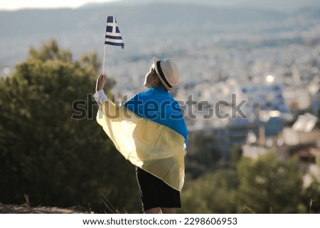 Smiling baby toddler, boy holding a Greece  and Ukrainian flag  on city background. Friendship country