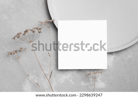 Blank square invitation card mockup with dry flowers decor on grey concrete background, top view