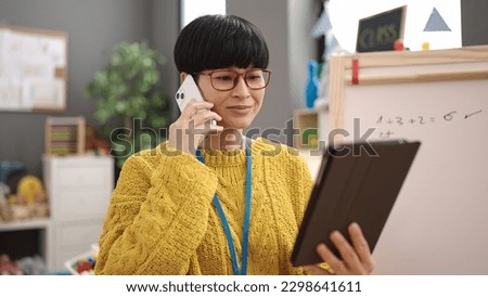 Young chinese woman preschool teacher talking on smartphone using touchpad at kindergarten