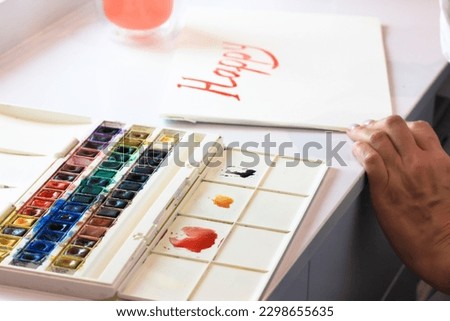 Artist painting with watercolor paints, writing with brush a word Happy. Creating DIY card for the happy New year. Paint palette on a white table. Drawing school, creating, ideas, hobby concept. 