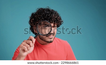 funny young latin spanish guy with a magnifying glass in his hands looks out or looks for something, dressed in casual, on a blue studio background. Royalty-Free Stock Photo #2298666861