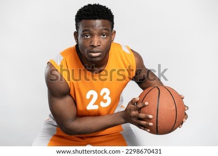 Portrait of young serious basketball player holding ball isolated on white background. Handsome African man wearing orange sportswear playing basketball looking at camera. Sport competition concept 