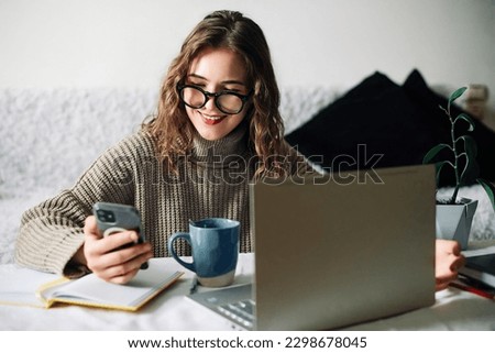 SMM specialist working from home at kitchen table, using smartphone to select blogger for product promotion in social media, in front of laptop. Influencer marketing concept Royalty-Free Stock Photo #2298678045
