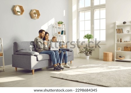 Happy family spend time in spacious room at home. Couple with child watch online movie on laptop sitting on sofa in cozy light living place space with white grey walls, shelves, house plants and wifi Royalty-Free Stock Photo #2298684345