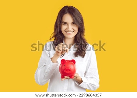 Portrait of satisfied happy woman who throws coin into piggy bank to save money. Beautiful smiling young Caucasian woman holding red piggy bank in shape of pig isolated on orange background.