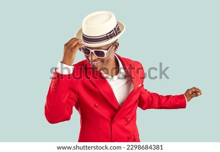 Time for party. Happy refined and stylish african man having fun dancing on pastel light blue background. Joyful positive man in red suit, white shirt, sunglasses holding on to hat having fun at party Royalty-Free Stock Photo #2298684381