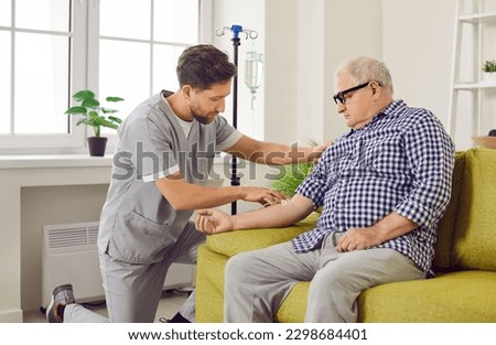 Nurse or doctor giving intravenous infusion to elderly patient. Young man in uniform inserts IV line needle in vein of old, retired man sitting on couch at home. Vitamin therapy, senior health concept Royalty-Free Stock Photo #2298684401