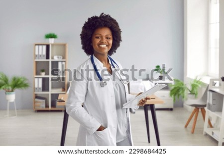 Portrait of African female doctor in lab coat standing in office. Beautiful smiling professional medical specialist with stethoscope around neck standing with clipboard and smiling looking at camera Royalty-Free Stock Photo #2298684425