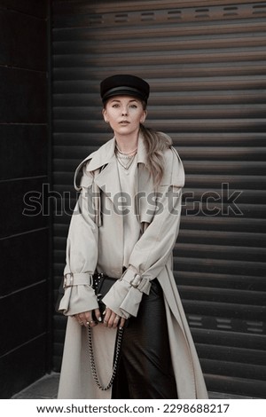 portrait of beautiful blonde girl dressed in beige trench coat, black leather midi skirt with slit, sweatshirt, cap, bag in hand, stylish fashion outfit, lifestyle model