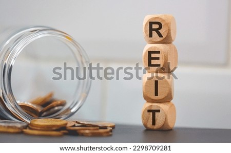 The acronym REIT for Real Estate Investment Trust written on wooden dice. A glass jar with coins in the composition.
