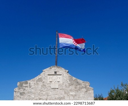 You see a flag of croatia near the village Nin. The colors of the flag are red, white and blue. It is a sunny day with a deep blue sky.