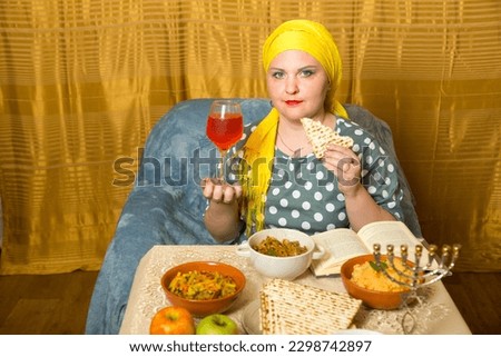At the festive table on Shabbat, a Jewish woman in a Kisui Rosh headdress eats matzah after blessing. Horizontal photo.