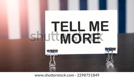 TELL ME MORE sign on paper on dark desk in sunlight. Blue and white background