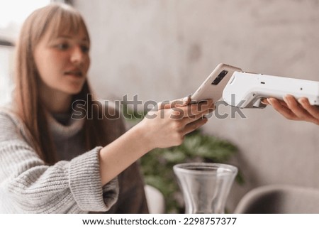 Blonde attractive woman paying with cellphone resting in cafe while waitress holding payment terminal. Girl relaxing in restaurant. Focus on part payment terminal and mobile phone. Royalty-Free Stock Photo #2298757377