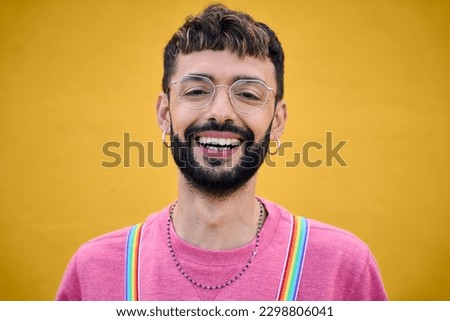 Close up smiling cheerful young Caucasian man holding his rainbow lgbt suspenders. Gay people posing for photo yellow background. Cheerful gay bearded person happy studio photo isolated. Millennials. Royalty-Free Stock Photo #2298806041