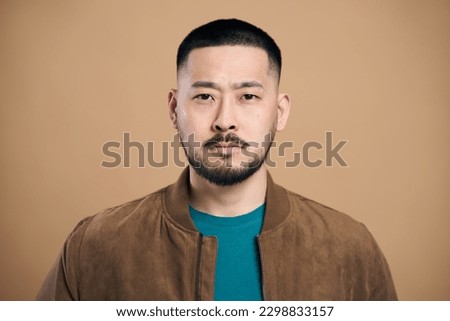 Portrait of self confident bearded asian man looking at camera with serious expression, unsmiling determined business man. Indoor studio shot isolated on brown background  Royalty-Free Stock Photo #2298833157