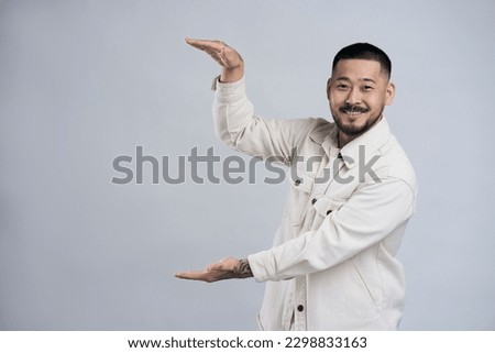 Portrait of smiling asian unshaven man showing advertisement area between his arms, copy space, big size. Indoor studio shot isolated on grey background 