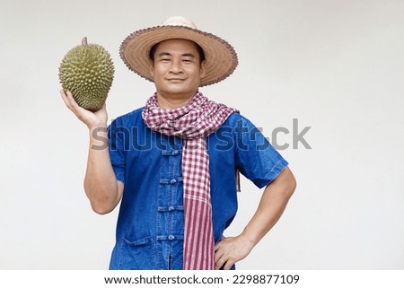 Handsome Asian man farmer wears hat, blue shirt, Thai loincloth scarf, holds durian fruit, feels confident. Concept, agriculture occupation. Thai farmers grow durian fruits as economic and export. 