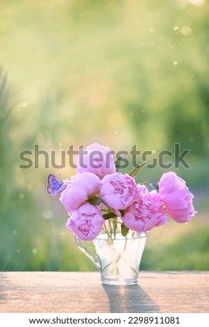 butterfly and pink peony flowers in glass cup on table in garden, abstract natural background. gentle romantic composition with flowers. spring, summer season. template for design. copy space