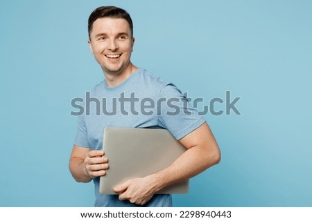 Young cheerful smiling happy IT man wearing casual t-shirt hold closed laptop pc computer looking aside on area isolated on plain pastel light blue cyan background studio portrait. Lifestyle concept