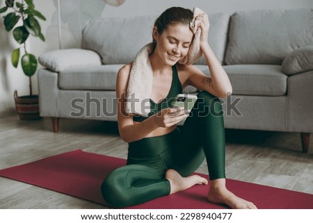 Full body happy young strong sporty athletic fitness trainer instructor woman wear green tracksuit towel use mobile cell phone sit on floor train do exercises at home gym indoor. Workout sport concept