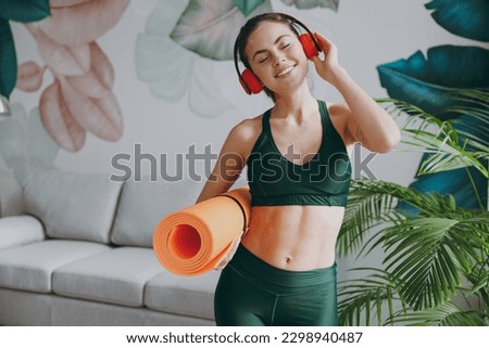 Young calm strong sporty athletic fitness trainer instructor woman wear green tracksuit headphones listen music hold in hand yoga mat training do exercises at home gym indoor. Workout sport concept Royalty-Free Stock Photo #2298940487