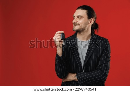 Young cool happy smiling fun successful unshaven latin man 20s wearing black striped jacket grey shirt holding car key fob keyless system looking aside isolated on red color background studio portrait