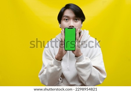 Happy stylish asian man holding phone and showing its green screen. Asian man in beige hoodie raising fist showing his screen phone with green screen template best for advertisment.