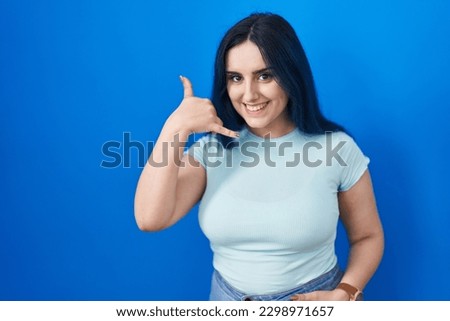 Young modern girl with blue hair standing over blue background smiling doing phone gesture with hand and fingers like talking on the telephone. communicating concepts. 