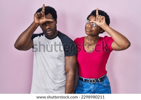 Young african american couple standing over pink background making fun of people with fingers on forehead doing loser gesture mocking and insulting. 