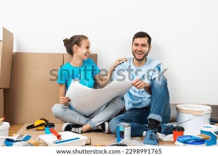 Young man and woman together planning their home renovation. Cardboard boxes, painting tools and materials on floor. House remodeling and interior renovation. People looking at blueprint at home.