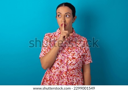 beautiful woman wearing floral dress over blue studio background makes silence gesture, keeps index finger to lips makes hush sign. Asks not to share secret.