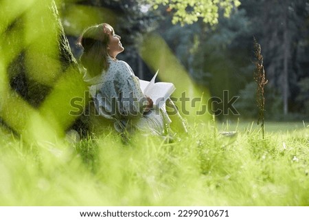 A young woman sits in a park leaning against a tree, reading a book in spring or summer, relaxed and happy, enjoying the sun. Royalty-Free Stock Photo #2299010671