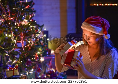 Christmastime, an expressive young girl with a santa hat opening her gift-box and finding a wonderful surprise in front of the christmas tree illuminated, in the warm atmosphere of christmas night