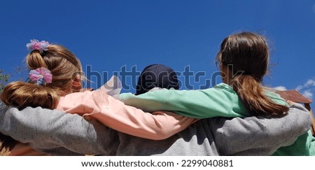 Three teenagers: two girls and a boy between them, hugging their shoulders, in the sun in the blue sky in spring (macro, rear view from the back). Royalty-Free Stock Photo #2299040881