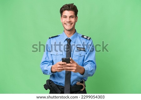 Young police man over isolated background sending a message with the mobile Royalty-Free Stock Photo #2299054303