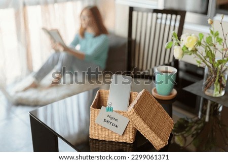 Smartphone is in separate wicker box with inscription digital detox on table. Woman reading book in background. Stop using digital gadgets. Mental and digital detox concept Royalty-Free Stock Photo #2299061371