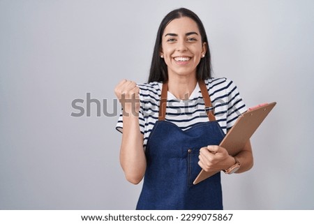 Young brunette woman wearing professional waitress apron and clipboard screaming proud, celebrating victory and success very excited with raised arms  Royalty-Free Stock Photo #2299075867
