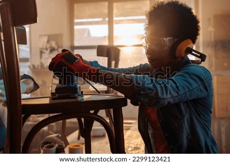 A crafty female African American carpenter is in a wood workshop using power tools to sand a chair. Royalty-Free Stock Photo #2299123421